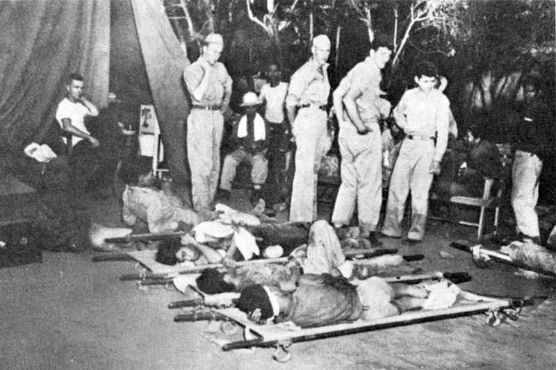 Photo of wounded men laying on stretchers on the floor with soldiers standing around them in a tent