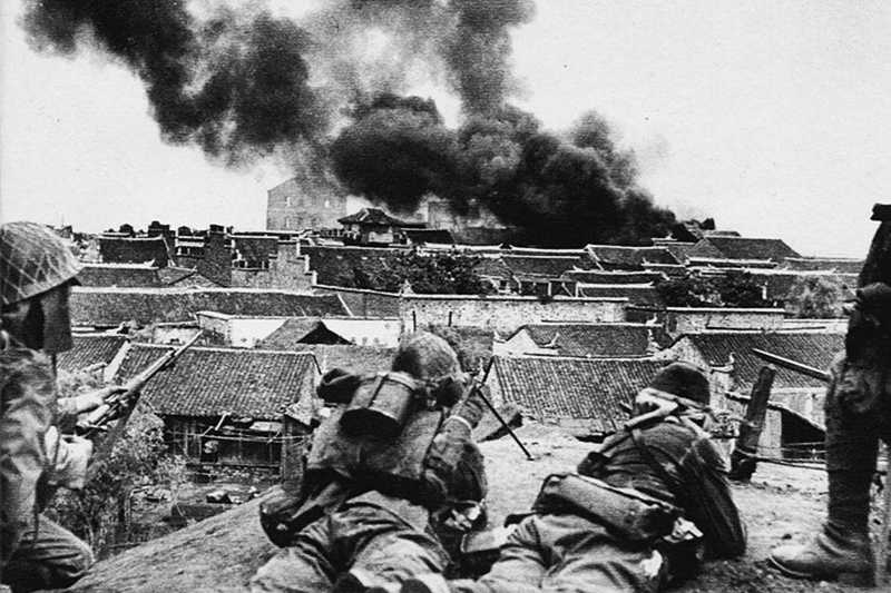 Black and white photo of Japanese soldiers laying on roof overlooking burning buildings