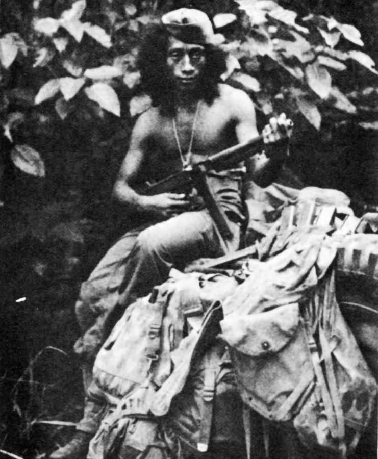 Photo of Filipino guerrilla holding a rifle, sitting on a jeep tire in the jungle