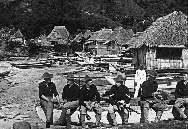 Black and white photo of American Soldiers seated on boat in a Filipino village