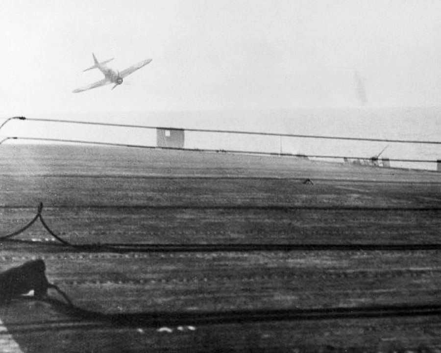 Photo looking over the deck of a ship with a plane in the distance heading towards it.