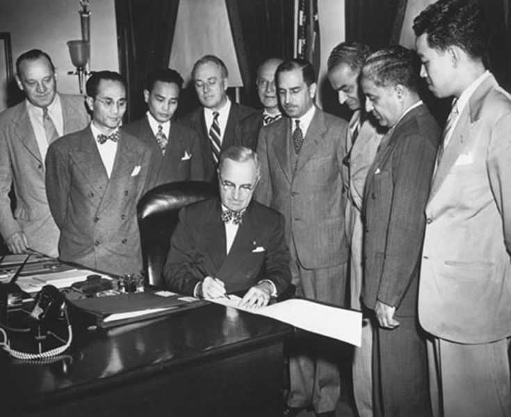 Photo of Harry Truman signing a document at his desk while several men stand around to watch