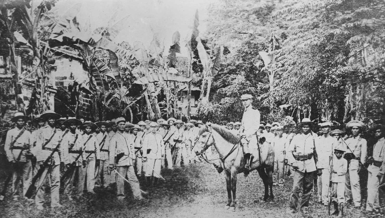 Black and white photo of Filipino soldiers in uniform and holding rifles in the jungle, with one man seated on a horse.